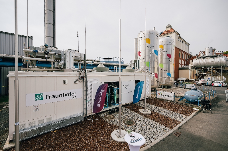 Versuchsanlage mit Containern und Tanks im Hintergrund, Firmenlogos und Beachflags Fraunhofer IEG und Stadtwerke Zittau