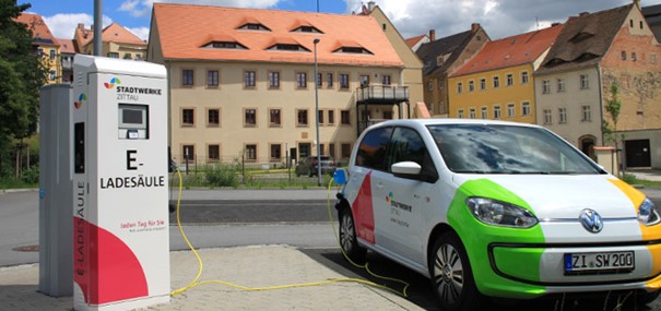 Ladesäule mit Stadtwerke Zittau Logo, rechts daneben steht ein Fahrzeug mit Stadtwerke Zittau Logo, mit angestecktem Ladekabel, vor einer Häuserfront